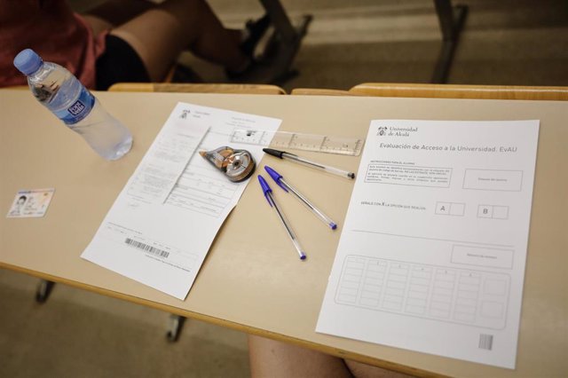 Vista de un examen, una botella de agua y bolígrafos en un pupitre mientras estudiantes de las asignaturas troncales de la rama de Ciencias 