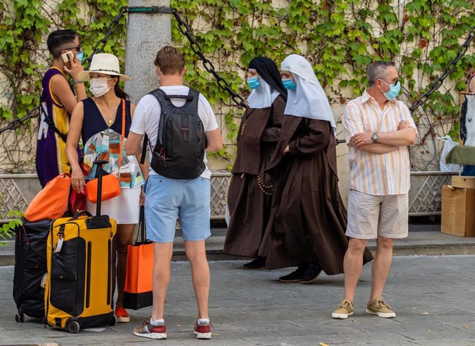 Unos turistas esperan el autobús mientras una pareja de monjas de la Orden de Santa Ángela de la Cruz  pasan por detrás con mascarillas en Sevilla