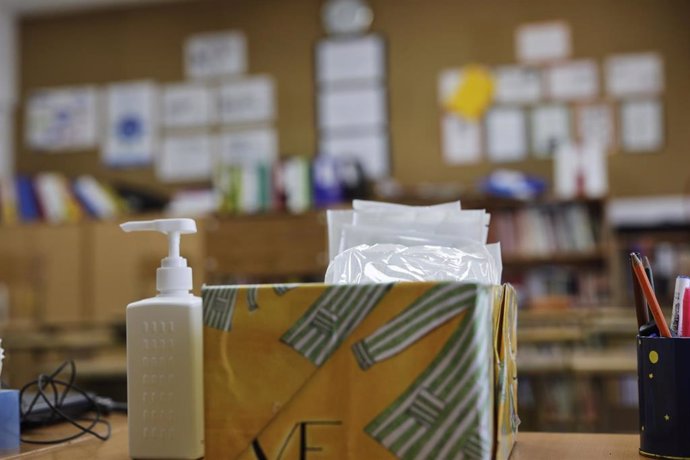 Mascarillas y gel desinfectante en la mesa del profesor de un aula donde se pueden observar los pupitres colocados y separados entre sí para el nuevo curso, foto de archivo