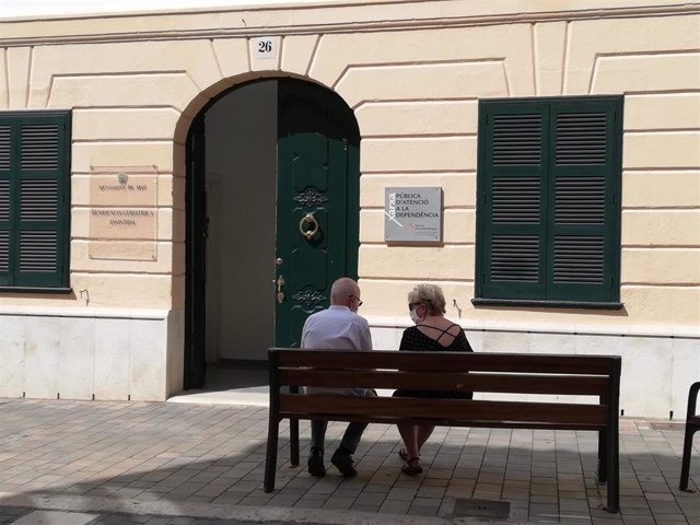 Dos personas mayores conversan sentadas en un banco y con mascarilla, ante la Residencia Geriátrica Asistida del Ayuntamiento de Mahón en la Calle Cós de Gràcia.