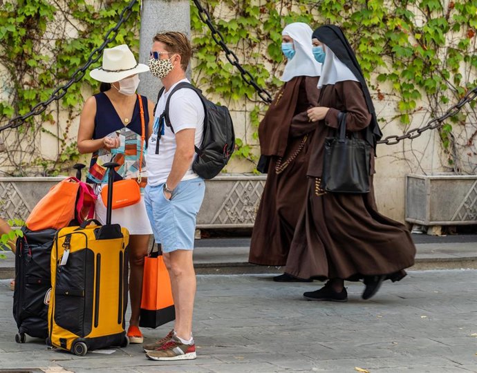 Unos turistas esperan el autobús mientras una pareja de monjas de la Orden de Santa Ángela de la Cruz  pasan por detrás con mascarillas el día que se ha anunciado la caída de un 98,9% de turistas extranjeros en Andalucía. Sevilla a 3 de agosto del 2020