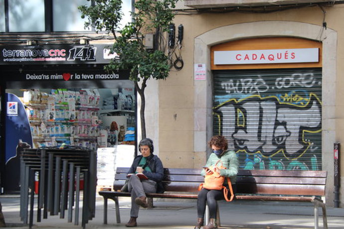 Pla obert de dues dones assegudes en un banc a la plaa de la Vila de Grcia, davant un resturant tancat per les noves restriccions. Imatge del 17 d'octubre de 2020. (Horitzontal)