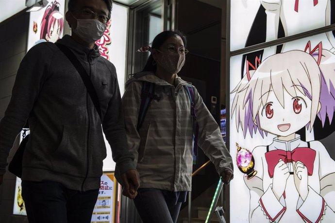 Dos personas con mascarilla pasan junto al escaparate de un local de juegos de targaperras pachinko decorado con un personaje de anime en Tokio, Japón