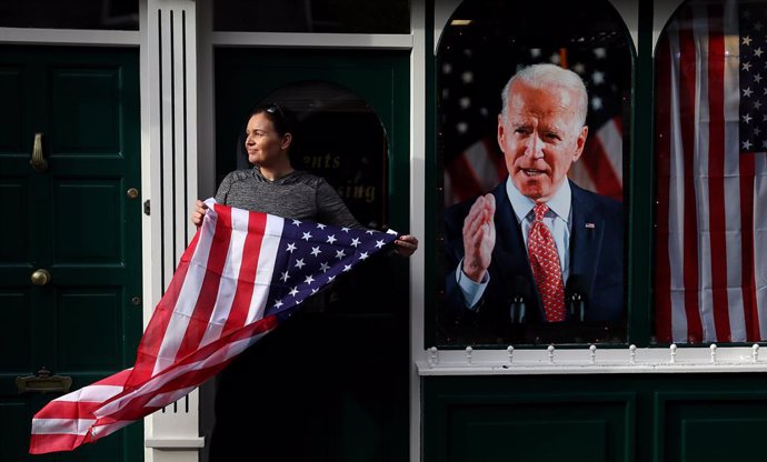 07 November 2020, Ireland, Ballina: Catherine Hallahan, owner of Hallihan's barbers, in Ballina holds the us national flag  as residents begin celebrations in the ancestral home of US Presidential candidate Joe Biden in anticipation of the results of th