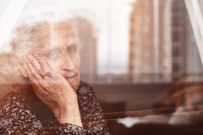 Mujer mayor sentada sola y triste tras una ventana.