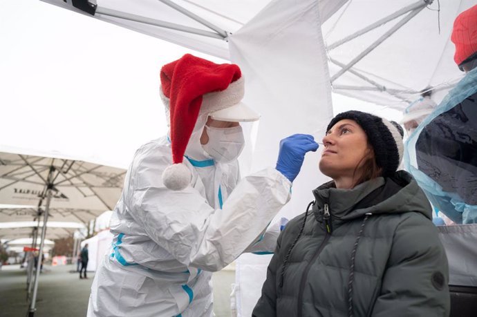 24 December 2020, Baden-Wuerttemberg, Goeppingen: A volunteer of the German Red Cross (DRK) wearing a Santa Claus hat takes a swap from a woman for a Coronavirus rapid test at a testing station. Tens of thousands of people across Germany  rush to get te