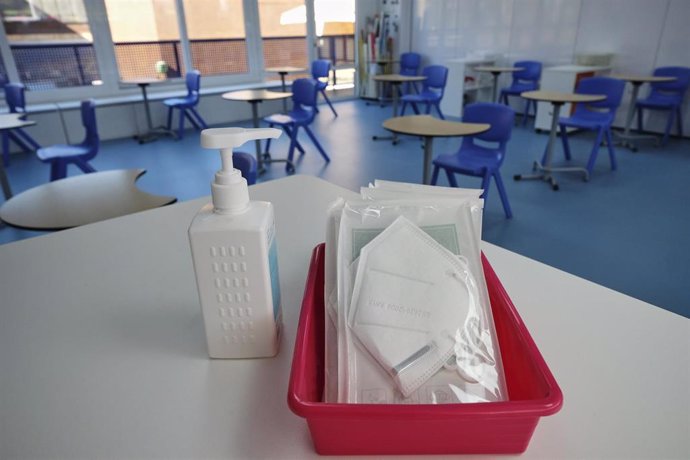 Mascarillas y gel desinfectante en la mesa del profesor de un aula donde se pueden observar los pupitres colocados y separados entre sí. 