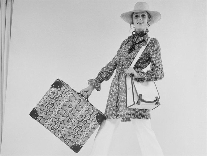 JJ models a pair of white trousers and hat with a suitcase in a reptile print, UK, 29th June 1970