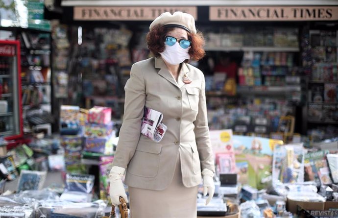 Una mujer protegida con mascarilla y con una revista en su brazo posa al lado de un quiosco.