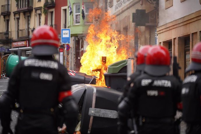 Varios agentes de la Ertzaintza observan como se quema un contenedor durante los altercados producidos en una manifestación contra el encarcelamiento de Pablo Hasel en Bilbao, Vizcaya, País Vasco (España), a 21 de febrero de 2021. 