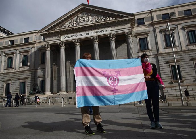 Dos personas sostienen una bandera trans durante una concentración convocada frente al Congreso de los Diputados en Madrid (España), a 18 de febrero de 2021. La Federación Plataforma Trans, acompañada de más representantes de colectivos trans y de asoci