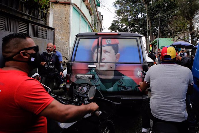 04 February 2021, Venezuela, Caracas: Scooter riders wait behind a vehicle marked with a photo of the late president and leader of the 1992 military uprising, Hugo Chavez during a march to commemorate the military uprising of 4 February 1992. Photo: Jes