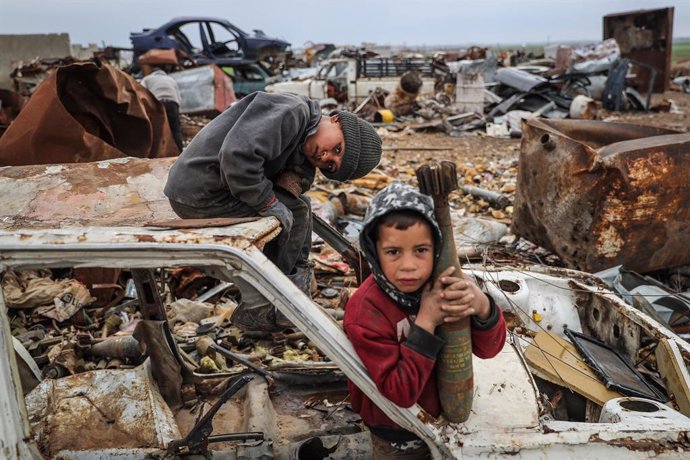 02 March 2021, Syria, Idlib: Malik Junaid (L), 9-years-old, sits on top of a destroyed car next to the 7-years-old Abdel Karim Hassan who carries a mortar shell, at a centre run by Junaid family for selling remnants of shellings and unexploded ordnances