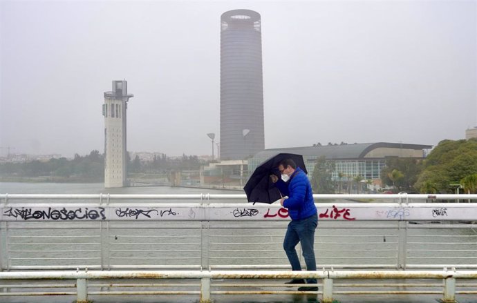 Un persona se protege de la lluvia y el viento caminando por la pasarela  peatonal de la Cartuja en Sevilla