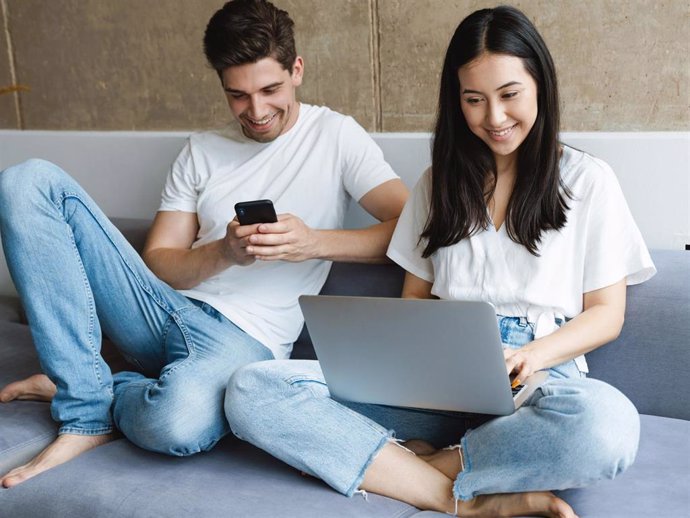 Archivo - Dos jóvenes utilizando un teléfono móvil y un ordenador portátil