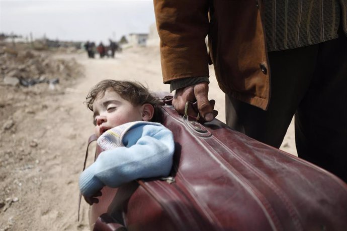 Archivo - Un hombre transporta a un niño en una maleta caminando hacia Hamourieh, donde se abrió una salida de evacuación desde el este de Ghouta.