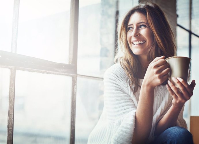 Archivo - Mujer sonriendo junto a una ventana con una taza.