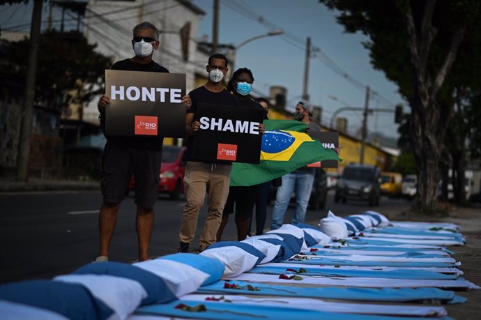 24 March 2021, Brazil, Acari: Demonstrators from the NGO Rio de Paz hold up placards outside Ronaldo Gazzola Hospital next to stretchers with sheets, pillows and roses during a protest in memory of the approximately 300,000 Coronavirus deaths in Brazil.