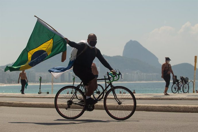 26 March 2021, Brazil, Rio De Janeiro: A supporter of Brazilian President Jair Bolsonaro rides a bicycle with the national flag at Copacabana during a protest against tightened Coronavirus restrictions in the city of Rio de Janeiro. Photo: Fernando Souz
