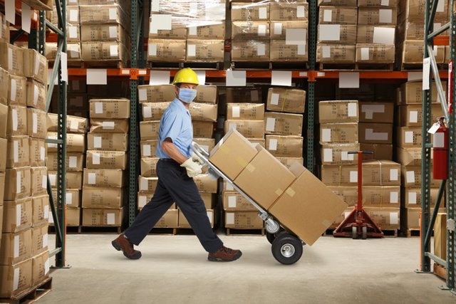 Archivo - Un trabajador del almacén con una máscara protectora y un casco empuja una carretilla de mano y una pila de cajas en un almacén lleno de inventario.