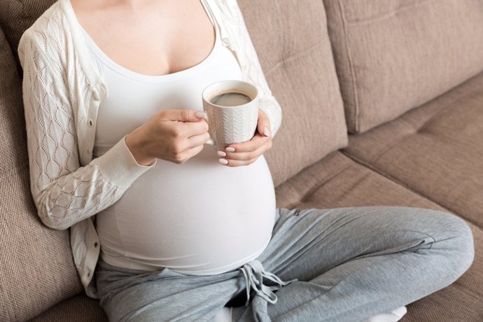 Archivo - Embarazada sentada en el sofá con café taza en café de la mano.