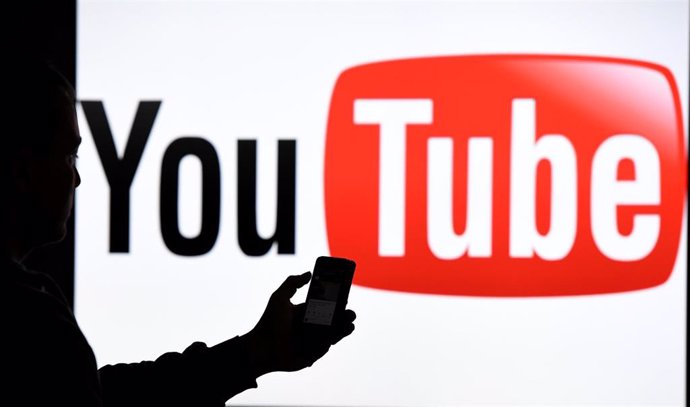 Archivo - Un hombre con un teléfono ante el logotipo de YouTube