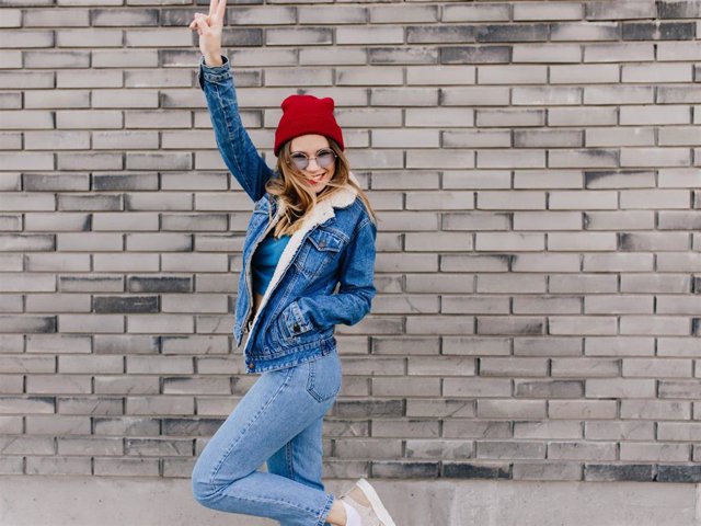 Archivo - Slim girl in trendy jeans having fun on the street in cold spring day. Blissful female model in denim attire dancing on urban background and waving hands.