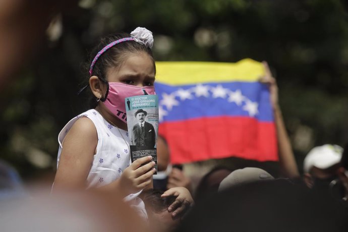 30 April 2021, Venezuela, Caracas: A girl with face mask sitting on the shoulders of an adult holds up a picture of Venezuelan doctor Jose Gregorio Hernandez in front of the church where Hernandez will be beatified. Hernandez is revered in Venezuela as 