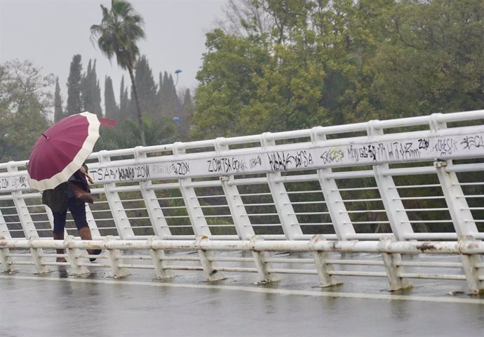 Archivo - Un persona se protege de la lluvia y el viento caminando por la pasarela  peatonal de la Cartuja en un día lluvioso y con fuertes rachas de viento en la provincia. En Sevilla (Andalucía, España), a 09 de febrero de 2021.
