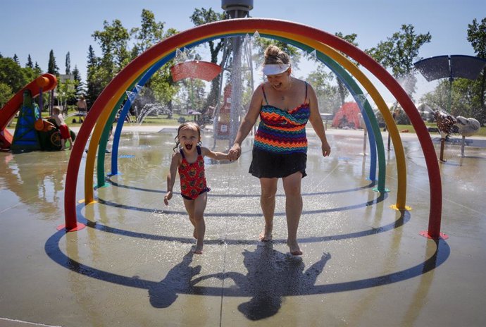 28 June 2021, Canada, Olds: Poppy Collins (L), five, and her mother Crystal Collins run through spray to try and beat the heat at a splash park. Environment Canada warns the torrid heat wave that has settled over much of Western Canada won't lift for da