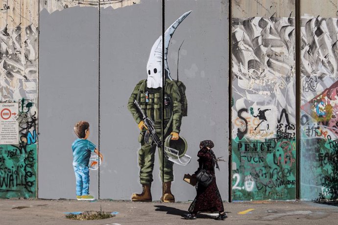 Archivo - 29 May 2021, Palestinian Territories, Bethlehem: A Palestinian woman walks past a graffiti mural depicting a boy holding a fishbowl looking at a uniformed Israeli soldier wearing a KKK-style hood, painted by Palestinian artist Taqi Spateen alo