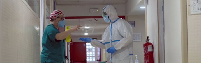 Un profesional de la sanidad con equipo de protección individual recibe ayuda de una compañera con producto de desinfección
