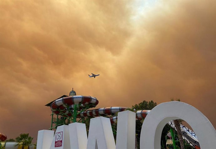 28 July 2021, Turkey, Side: Smoke drifts over leisure facilities at a hotel complex in the Turkish resort region of Antalya as a plane is on approach to land. Winds drove flames from several wildfires toward residential districts, the district administr