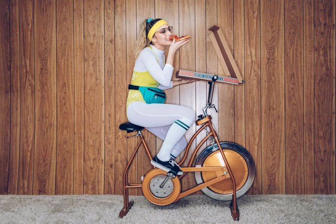 Archivo - Mujer comiendo una pizza mientras hace ejercicio en una bicicleta estática.