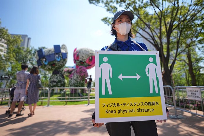 24 July 2021, Japan, Tokyo: A staff member holds a sign with a symbol and the words "Keep physical distance" to keep enough distance to protect themselves and others from contracting the coronavirus during the Tokyo 2020 Olympic Games. Visitors in the b