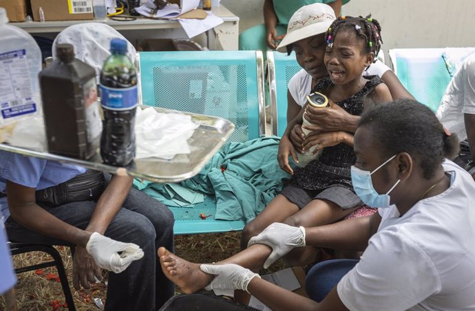 16 August 2021, Haiti, Les Cayes: A young girl is held by her mother as nurses care for a wound on her foot at OFATMA Hospital, as the patients afraid of hospital collapse after it's walls and floor cracked during the earthquake. Rescuers in Haiti are w