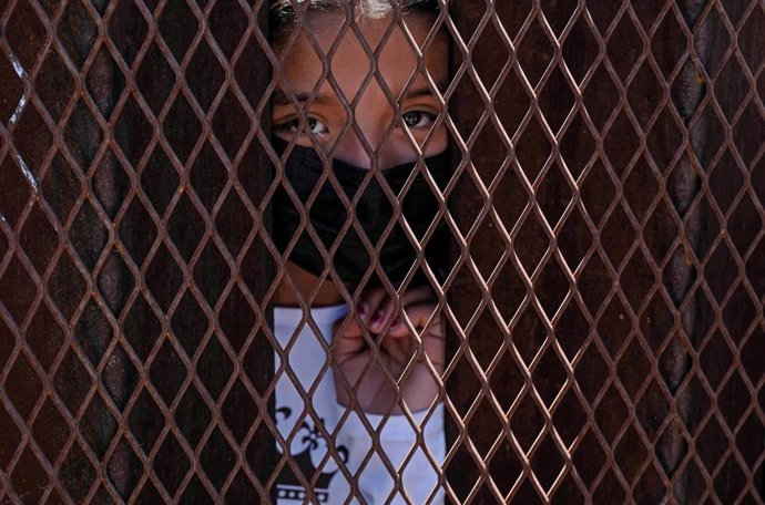 Archivo - Un joven migrante solicitante de asilo se encuentra detrás de la valla del muro fronterizo que separa Estados Unidos y México. 