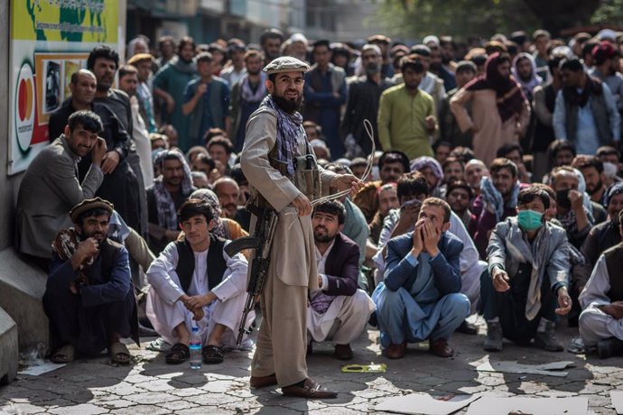 FILED - 22 September 2021, Afghanistan, Kabul: A Taliban security guard plays with a whip while standing in front of Afghan men waiting outside a bank to withdraw money. Photo: Oliver Weiken/dpa