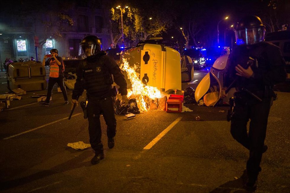 Varios mossos antidisturbios pasan delante de un contenedor quemado en la plaza Urquinaona durante una manifestación por el referéndum del 1-O en Catalunya, a 1 de octubre de 2021, en Barcelona