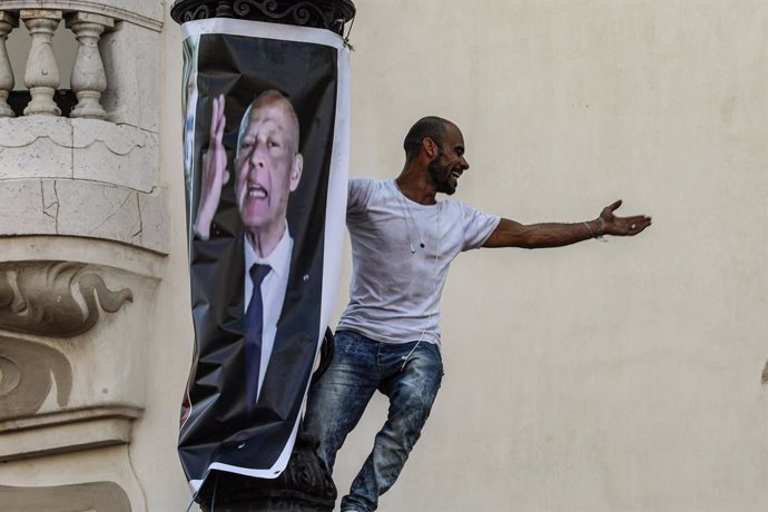 03 October 2021, Tunisia, Tunis: A man gestures as he climbs a light pole next to a poster bearing the image of Tunisian President Kais Saied during a protest supporting the latter, after he suspended parliament and assumed executive authority in July 2