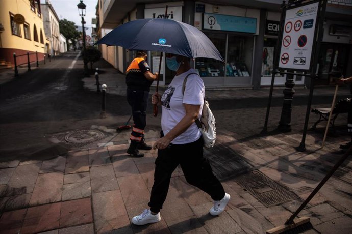 Una mujer se protege de la ceniza con un paraguas, mientras un agente de Protección Civil retira la ceniza de las calles del municipio de Los Llanos de Aridane