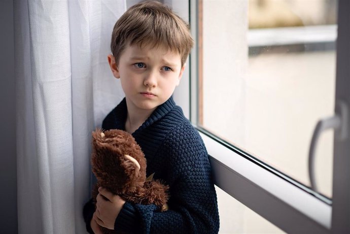 Archivo - Niño está abrazando a su osito de peluche. De pie junto a la ventana. Día lluvioso. Concepto de soledad y espera