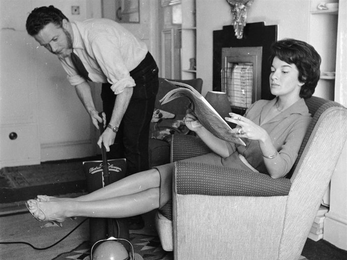 Archivo - An employee of the 'Housewives Help Service' hoovering the living room of a businesswoman while she relaxes.