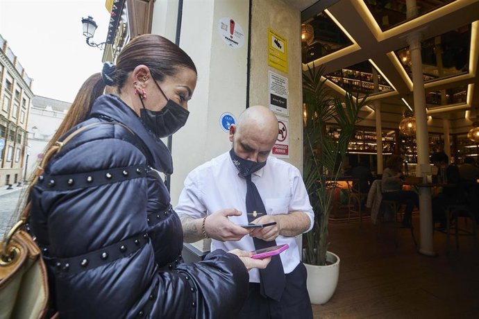 Un camarero escanea un certificado en la puerta de un bar a una clienta durante el primer día de petición de Certificados Covid-19 para la hostelería, a 20 de diciembre de 2021 en Sevilla 