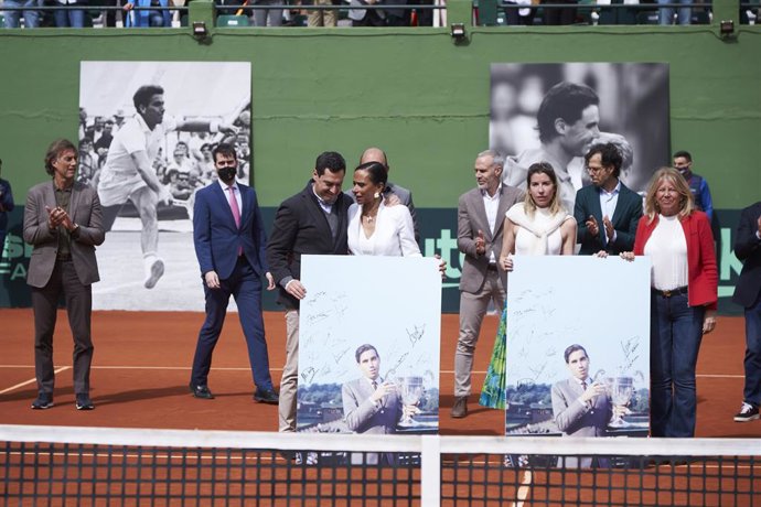 El presidente de la Junta, Juanma Moreno, entrega a la viuda de Manolo Santana, Claudia Inés, una foto de su difunto marido durante el homenaje a Manolo Santana en La ronda clasificatoria de la Copa Davis entre España y Rumania en el Club de Tenis Puent