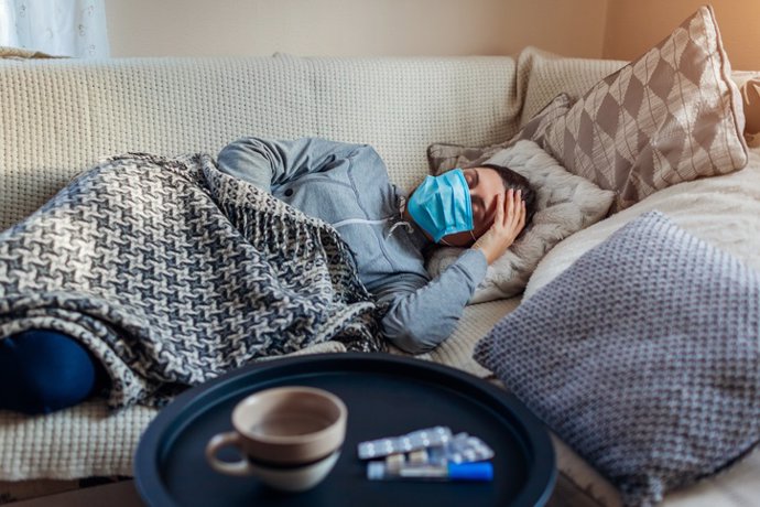 Archivo - Mujer enferma con gripe o resfriado. Chica acostada en la cama con dolor de cabeza con máscara protectora por pastillas y agua sobre la mesa.