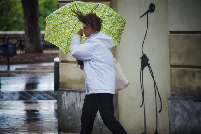 Archivo - Una mujer camina por la calle sujetando su paráguas, movido por el viento.