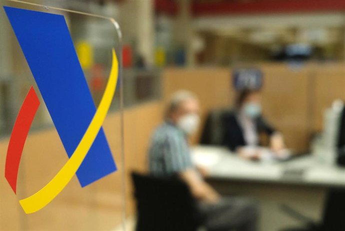 Archivo - Señal en un cristal del icono de la Agencia Tributaria mientras una empleada atiende a un hombre de manera presencial desde una oficina de la Agencia Tributaria, a 2 de junio de 2021