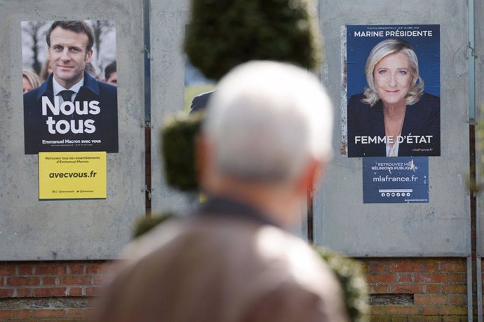 11 April 2022, France, Denain: A man walks past campaign posters of the French presidential candidates, incumbent President Macron (L) of the iberal party La Republique en Marche (LREM) and presidential candidate of the far-right Rassemblement National 