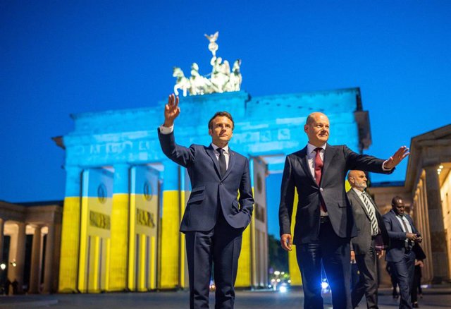 French President Emmanuel Macron and German Chancellor Olaf Scholz stand in front of the Brandenburg Gate in Berlin, which is illuminated in the colors of the Ukrainian flag.